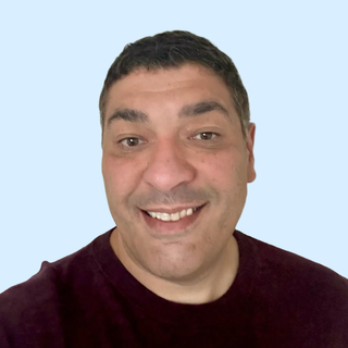 Smiling person with short dark hair wearing a dark shirt against a light blue background.