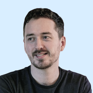 A man with short dark hair and a beard, wearing a dark shirt, smiles slightly against a light blue background.