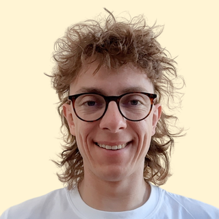 Smiling person with curly, tousled hair and glasses, wearing a white T-shirt, against a light background.