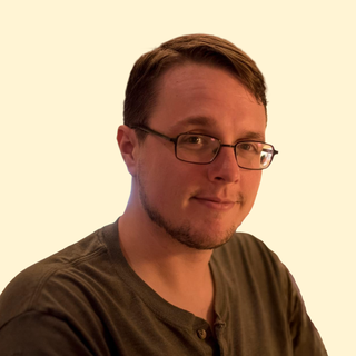 A person with glasses and a short beard smiles while wearing a dark henley shirt; the background is light in color.