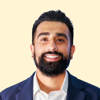 Smiling man with a beard, wearing a white shirt and blue blazer, against a light beige background.