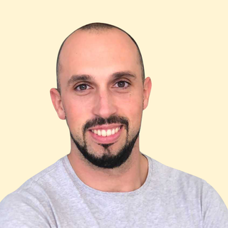 Smiling man with a beard and shaved head, wearing a light gray shirt, against a plain beige background.