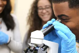 A student wearing blue gloves looks through a microscope while two other students in the background smile and watch in a science classroom.