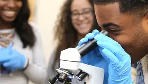 A student wearing blue gloves looks through a microscope while two other students in the background smile and watch in a science classroom.