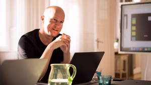 Person sitting at a desk in a bright office, smiling and gesturing while talking. Two laptops, a glass jug of water, and a few glasses are on the table, with a presentation slide visible on a screen in the background.