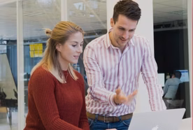 Two people in an office setting looking at a laptop screen together. One person is seated and typing, while the other stands beside them, gesturing toward the screen in discussion.