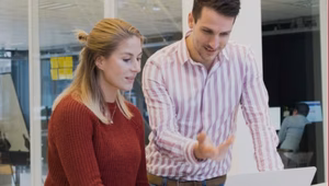 Two people in an office setting looking at a laptop screen together. One person is seated and typing, while the other stands beside them, gesturing toward the screen in discussion.