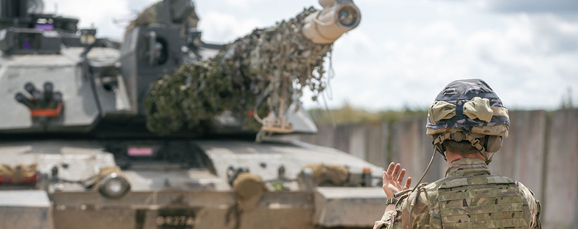 A soldier in camouflage gear guides a tank with a camouflaged barrel on a dirt road, under a cloudy sky.