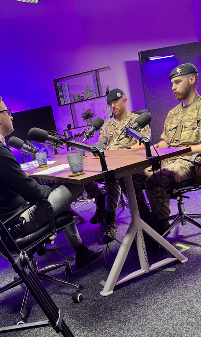 Three men, two in military uniforms, sit at a table with microphones in a studio with purple lighting, being recorded by a camera.