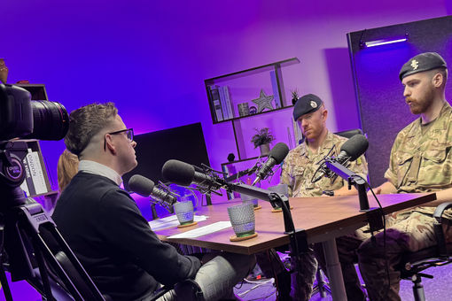 Three men, two in military uniforms, sit at a table with microphones in a studio with purple lighting, being recorded by a camera.