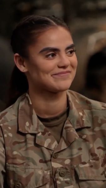 A woman in a camouflage military uniform smiles, seated with a blurred background.