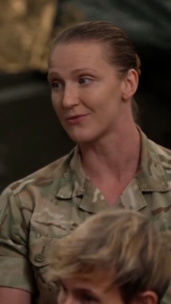 Person in military camouflage uniform speaking, with short hair, visible indoors.