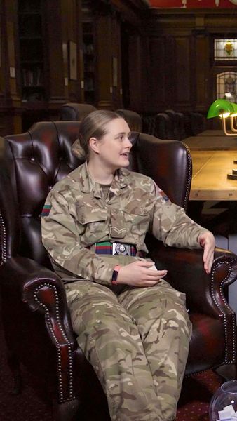 A person in military uniform sits in a leather armchair, in a room with wood paneling and a green lamp on a table in the background.