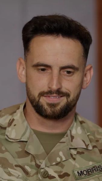 A person in a military camouflage uniform with a name tag, looking downward with a slight smile.