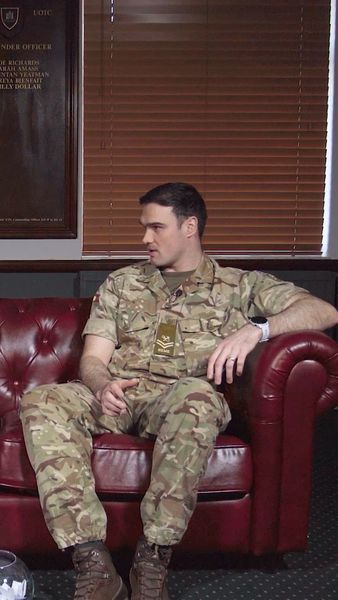 A person in camouflage military uniform sitting on a red leather couch, looking to the side, with a wooden plaque and blinds in the background.