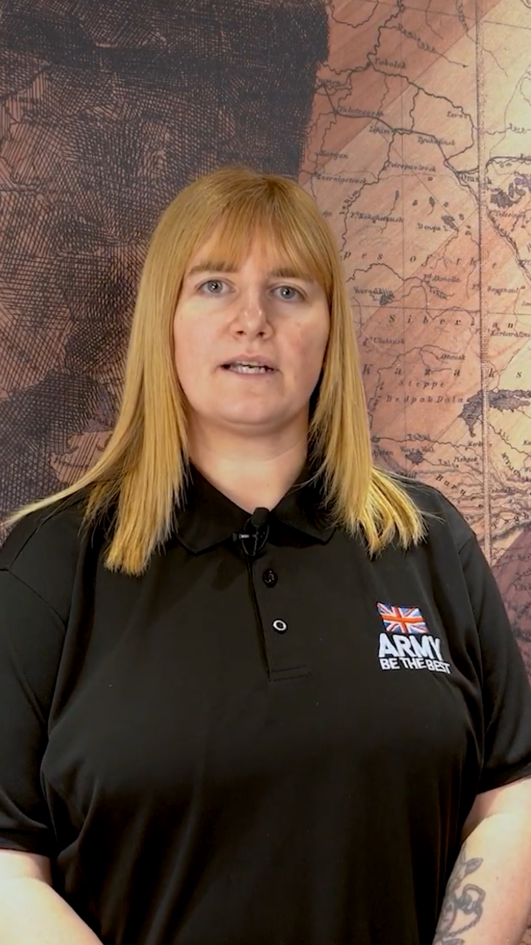A person with straight, light brown hair wearing a black polo shirt with a British Army logo stands in front of an illustrated map backdrop.