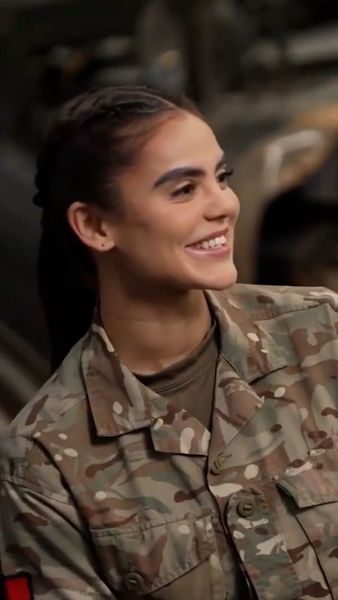 A smiling person in a camouflage military uniform with braided hair, standing outdoors.