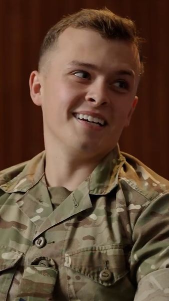 Smiling person in military camouflage uniform, looking to the side with short hair, sitting against a neutral brown background.