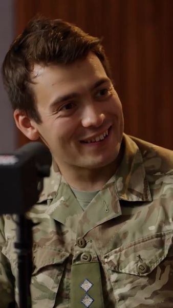 A smiling man in military camouflage uniform sits indoors, looking to the side, with a microphone in the foreground.
