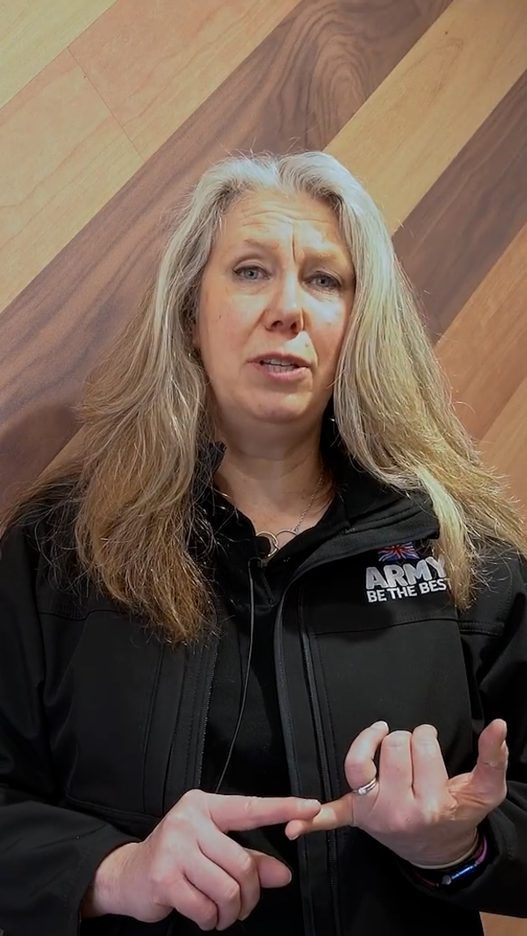 A woman with long hair wearing a black jacket talks with hand gestures against a wooden background. The jacket has "Army Be The Best" written on it.