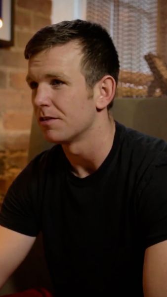 A person with short, dark hair wearing a black t-shirt, sitting indoors with a brick wall and curtains in the background.