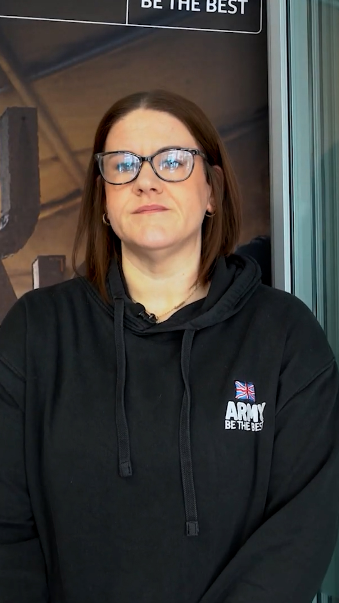 A woman wearing glasses and a black hoodie with "Army Be The Best" logo stands against a backdrop with similar text.