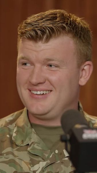 Smiling person in military uniform speaking into a microphone, with a wooden background.