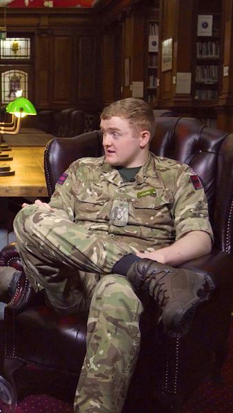 A person in military uniform sits cross-legged in a leather armchair in a dimly lit library, with bookshelves and green desk lamps in the background.