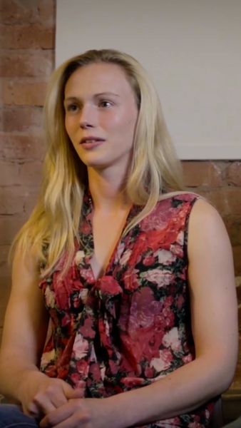 A person with long blonde hair wearing a red floral sleeveless top sits against a brick wall, looking slightly to the side.
