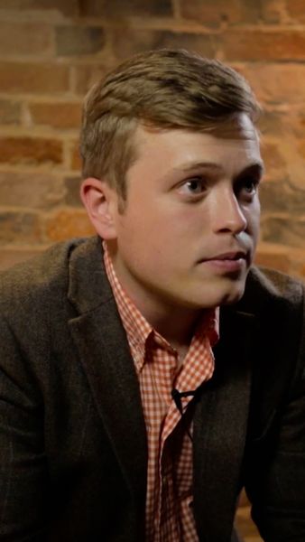 A man wearing a brown blazer and checkered shirt, sitting against a brick wall, looking to the right.