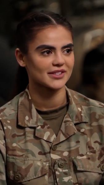 A person in a camouflage military uniform, smiling, with hair pulled back, facing slightly to the left.