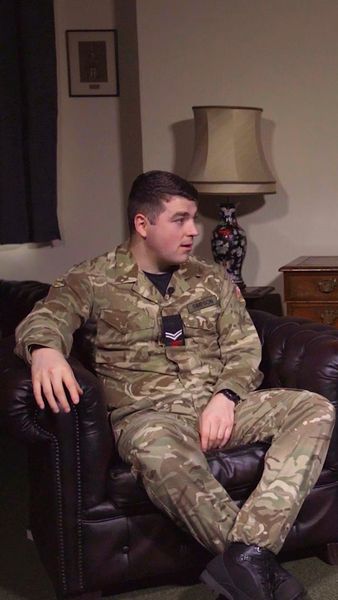 A person in camouflaged military attire sits on a brown leather couch in a room with a lamp, artwork, and wooden furniture.