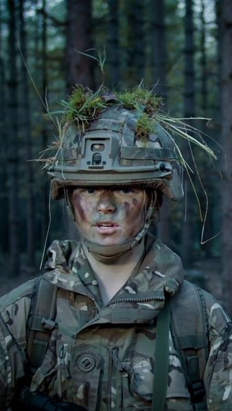 A soldier in camouflage gear and face paint stands in a forest, wearing a helmet adorned with grass and leaves for concealment.