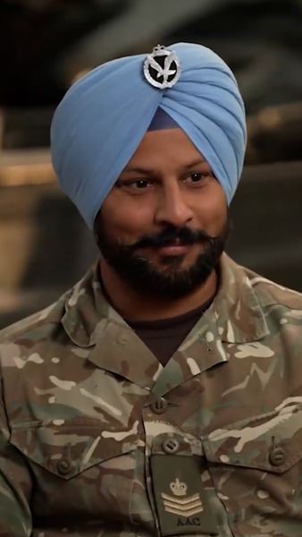 Man wearing a military uniform with a patterned fabric and a light blue turban adorned with an insignia, smiling gently.