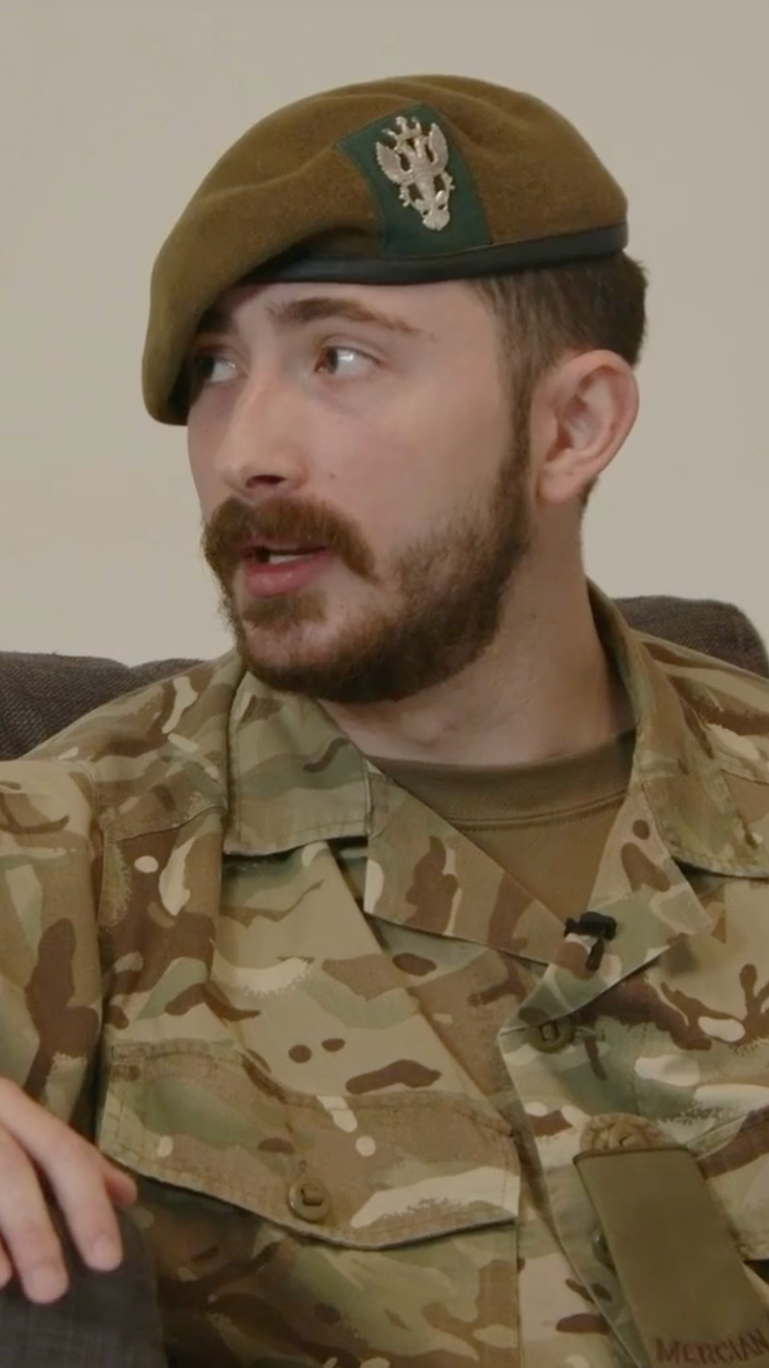 A man in camouflage military uniform and a beret looks to the side, sitting on a sofa.