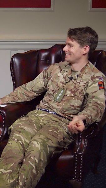 A person in military camouflage uniform sits in a leather armchair, smiling and gesturing slightly with their hand.