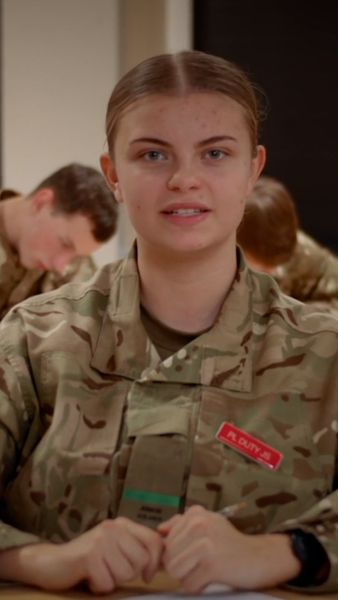 Person in military uniform with name tag, looking at the camera, seated at a table. Other uniformed individuals are in the background.
