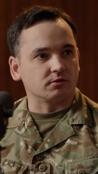 A person in a camouflage military uniform looks to the side, with a focused expression in an indoor setting.
