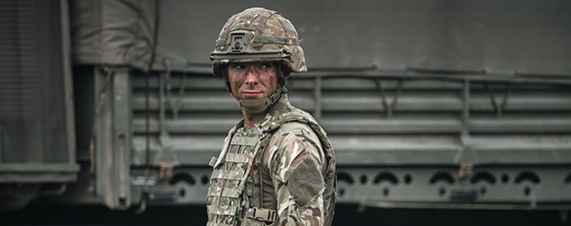 A soldier in camouflage uniform and helmet stands alert in front of a military vehicle, with como paint on his face.