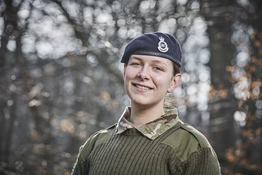Young person in military uniform and beret, smiling outdoors with blurred trees in the background.