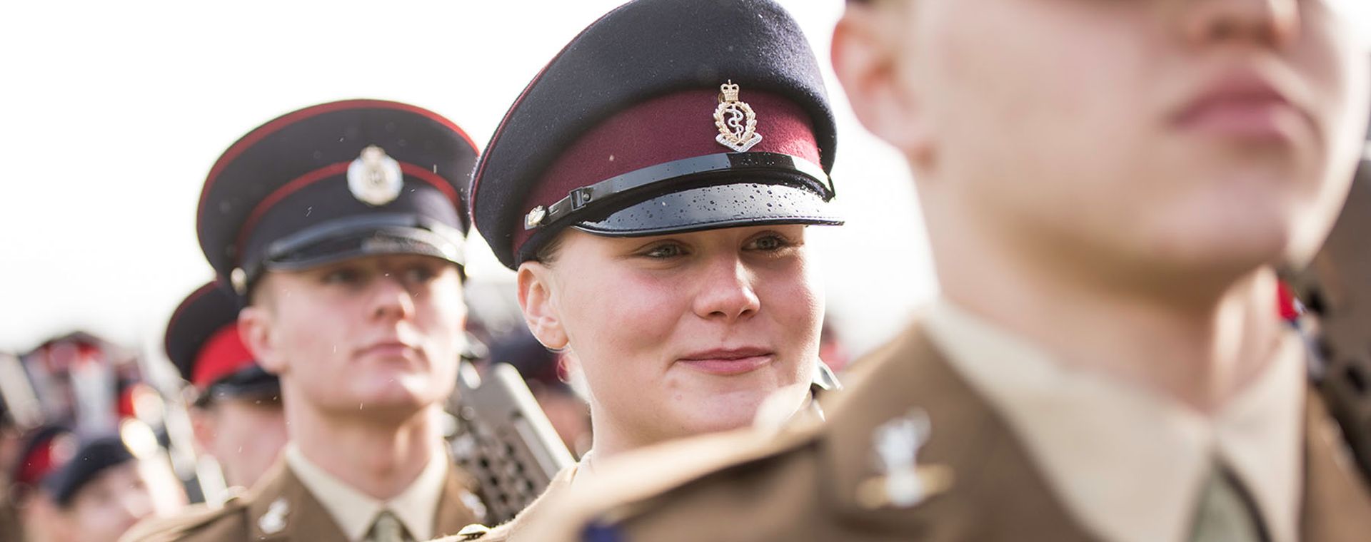 Soldiers in uniform stand in formation outdoors, wearing caps with insignia, focused expressions, and neatly arranged attire.