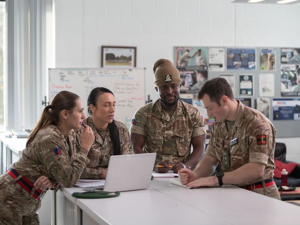 Four people in military uniforms gather around a table in an office, discussing documents and notes.