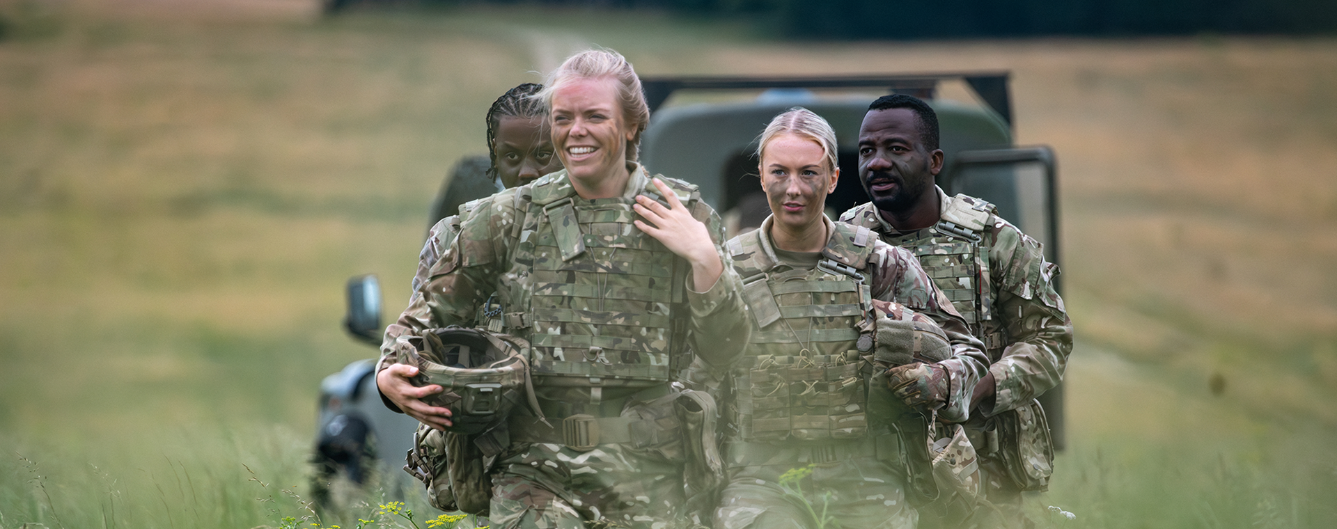 Four soldiers in camouflage uniforms walk through a grassy field, smiling, with a military vehicle in the background.
