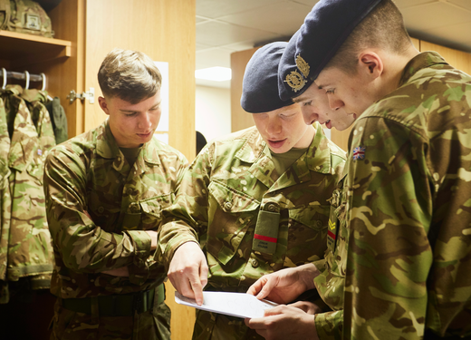 Three soldiers in camouflage uniforms review a document together in a room with military gear in the background.