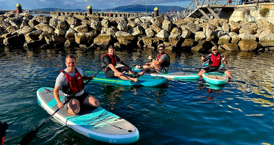 Four people wearing life vests sit on paddleboards in a calm harbor with rocky shores under a blue sky.