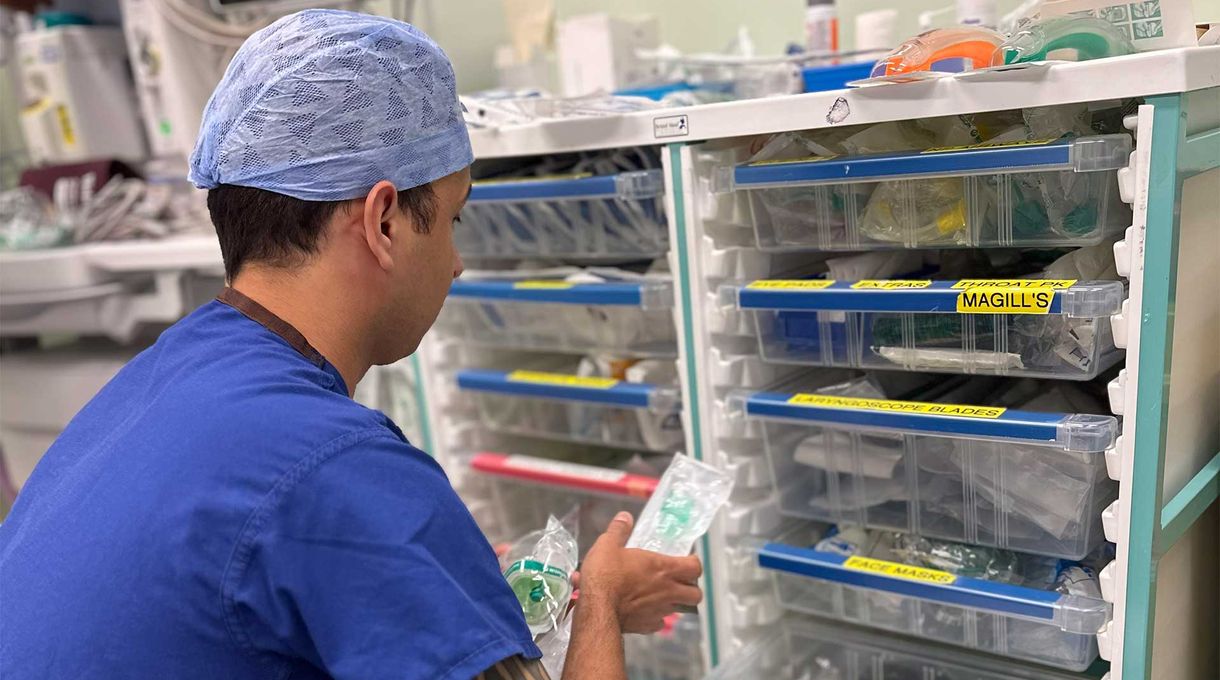 Medical professional in blue scrubs and cap organizing supplies in a hospital storage unit with labeled drawers.