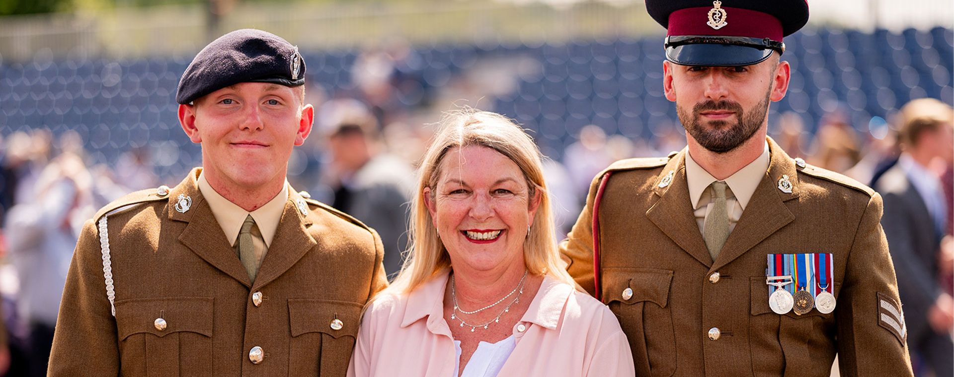 A smiling woman stands between two uniformed soldiers, one wearing a beret and the other a peaked cap with medals, at an outdoor event.