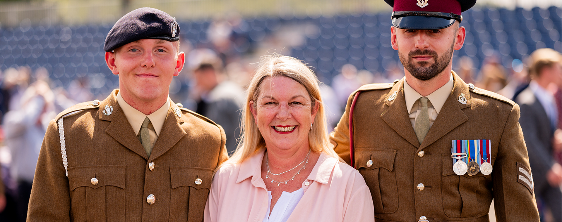 A smiling woman stands between two uniformed soldiers, one wearing a beret and the other a peaked cap with medals, at an outdoor event.