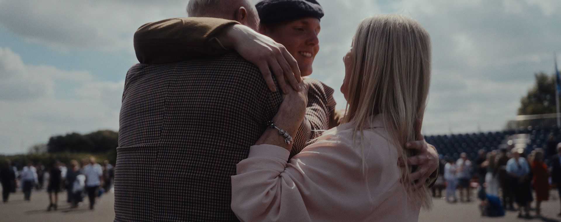 A smiling young soldier in a beret hugs two adults tightly, surrounded by a crowd and cloudy sky in the background.