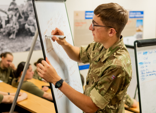 A military instructor writing on a whiteboard in a classroom, with soldiers seated at desks focusing on the lesson.
